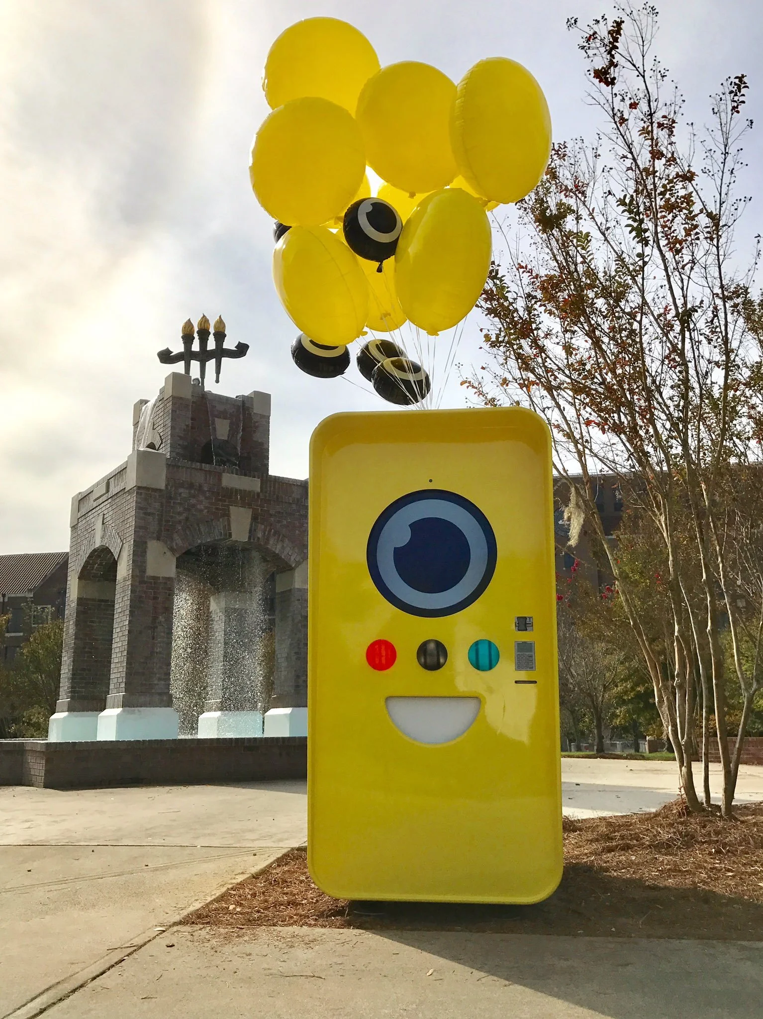 Yellow Snapbot Spectacles vending machine with balloons beside a brick fountain and torch monument on a campus plaza.