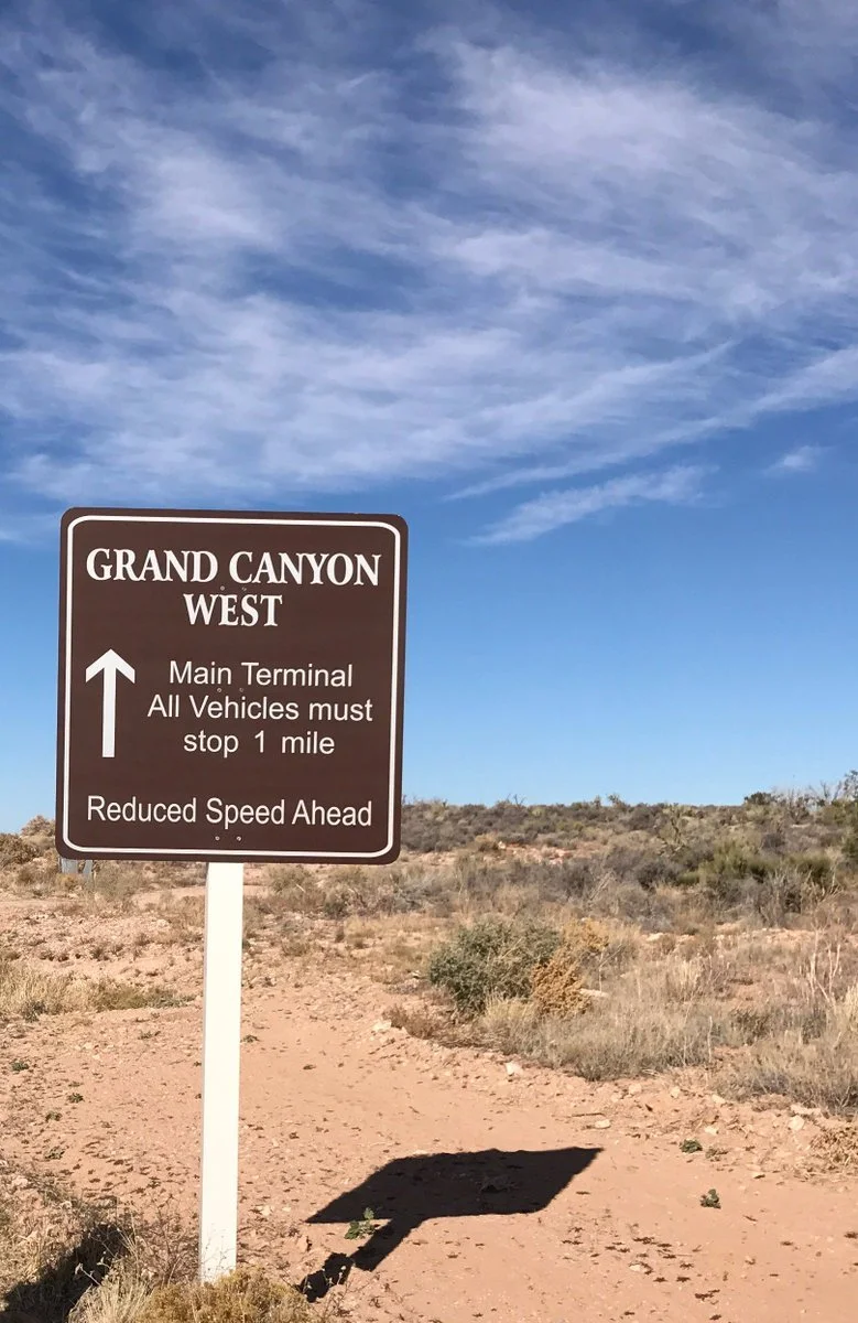 Brown road sign reading "Grand Canyon West, Main Terminal, All Vehicles must stop 1 mile, Reduced Speed Ahead" in desert.