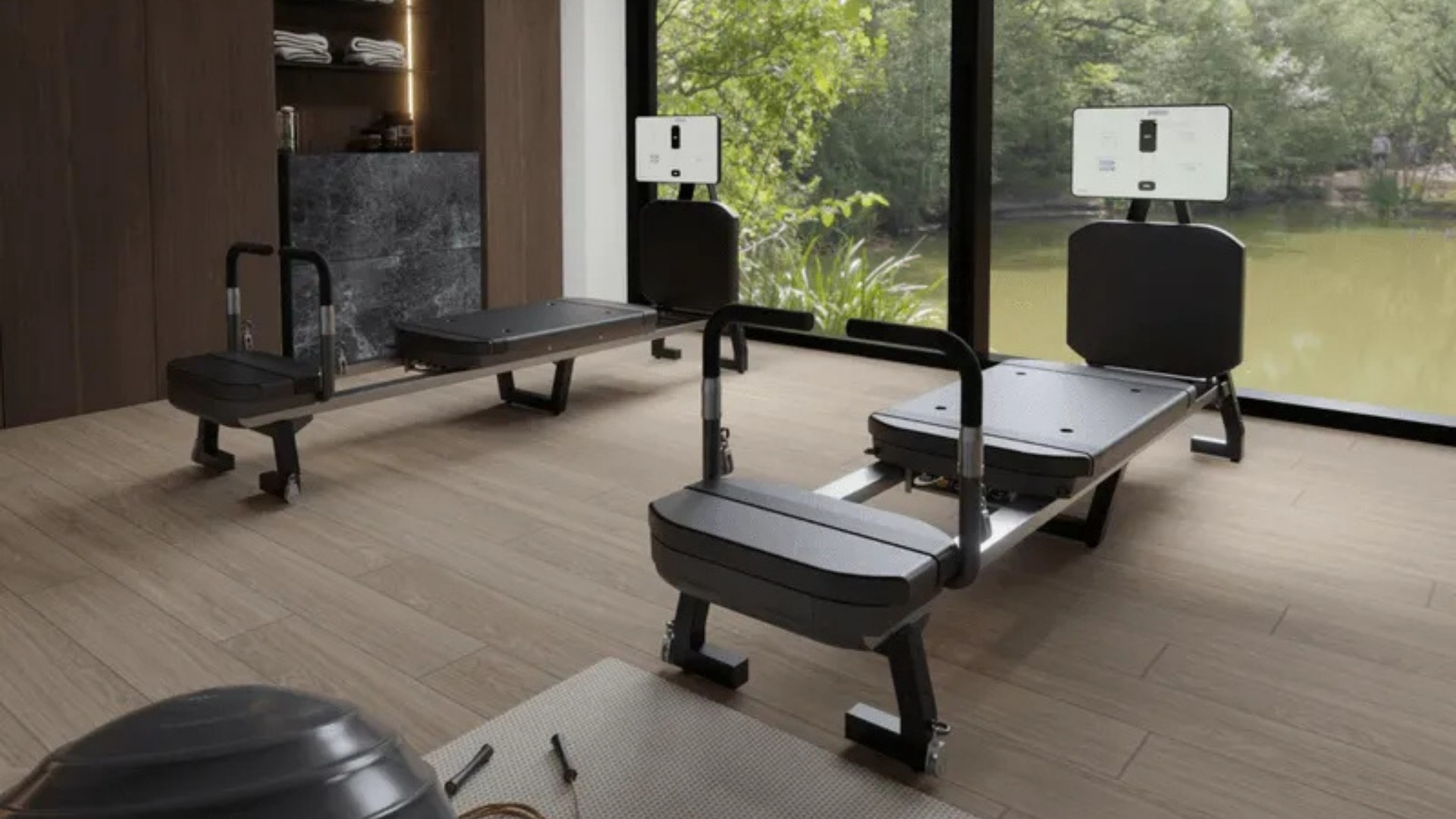 Two black Skop reformer machines with screens in a wood-floored home gym beside a floor-to-ceiling window facing a pond.