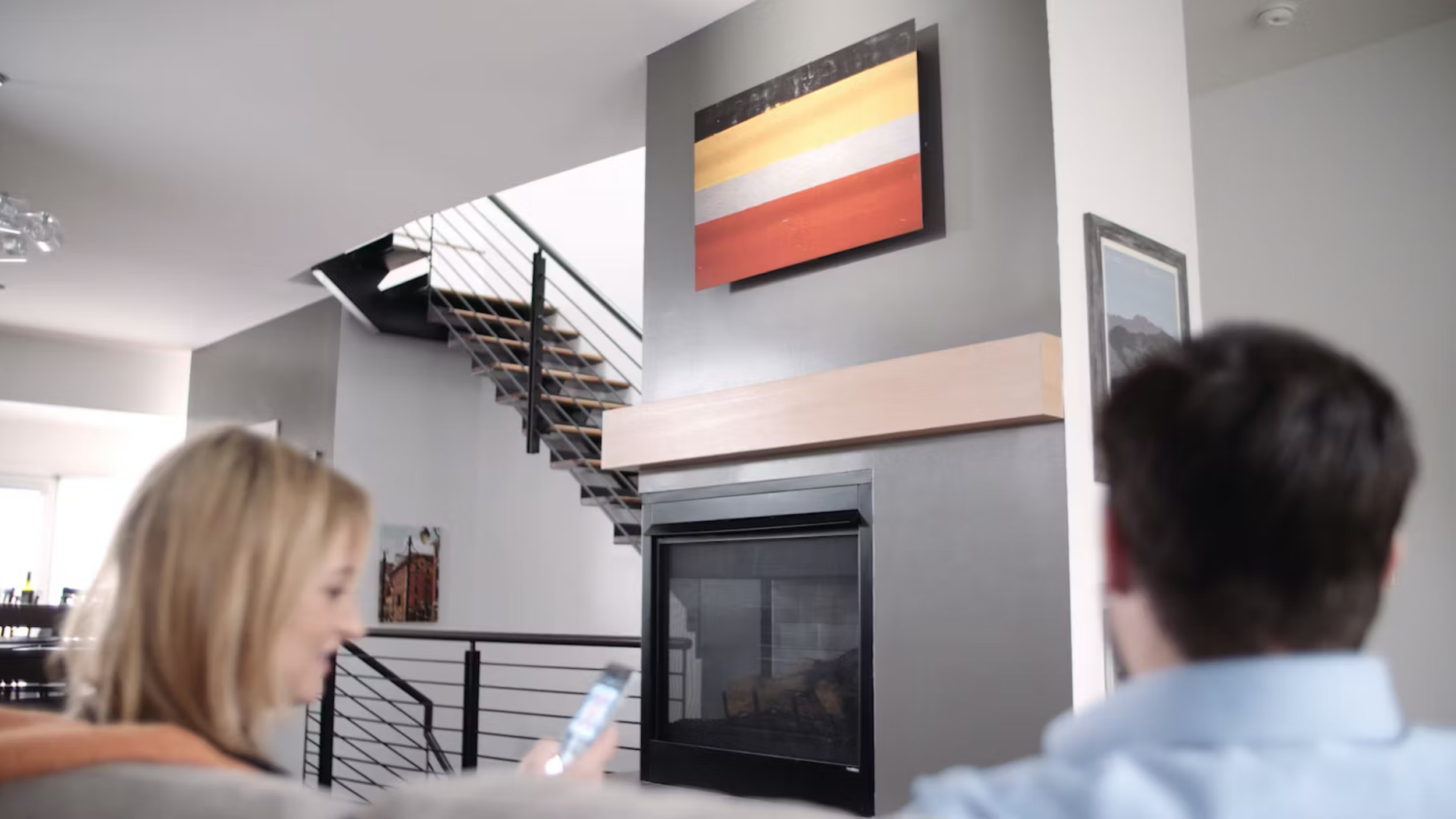 Living room with a striped orange, white and yellow canvas above a fireplace as two people sit with a phone