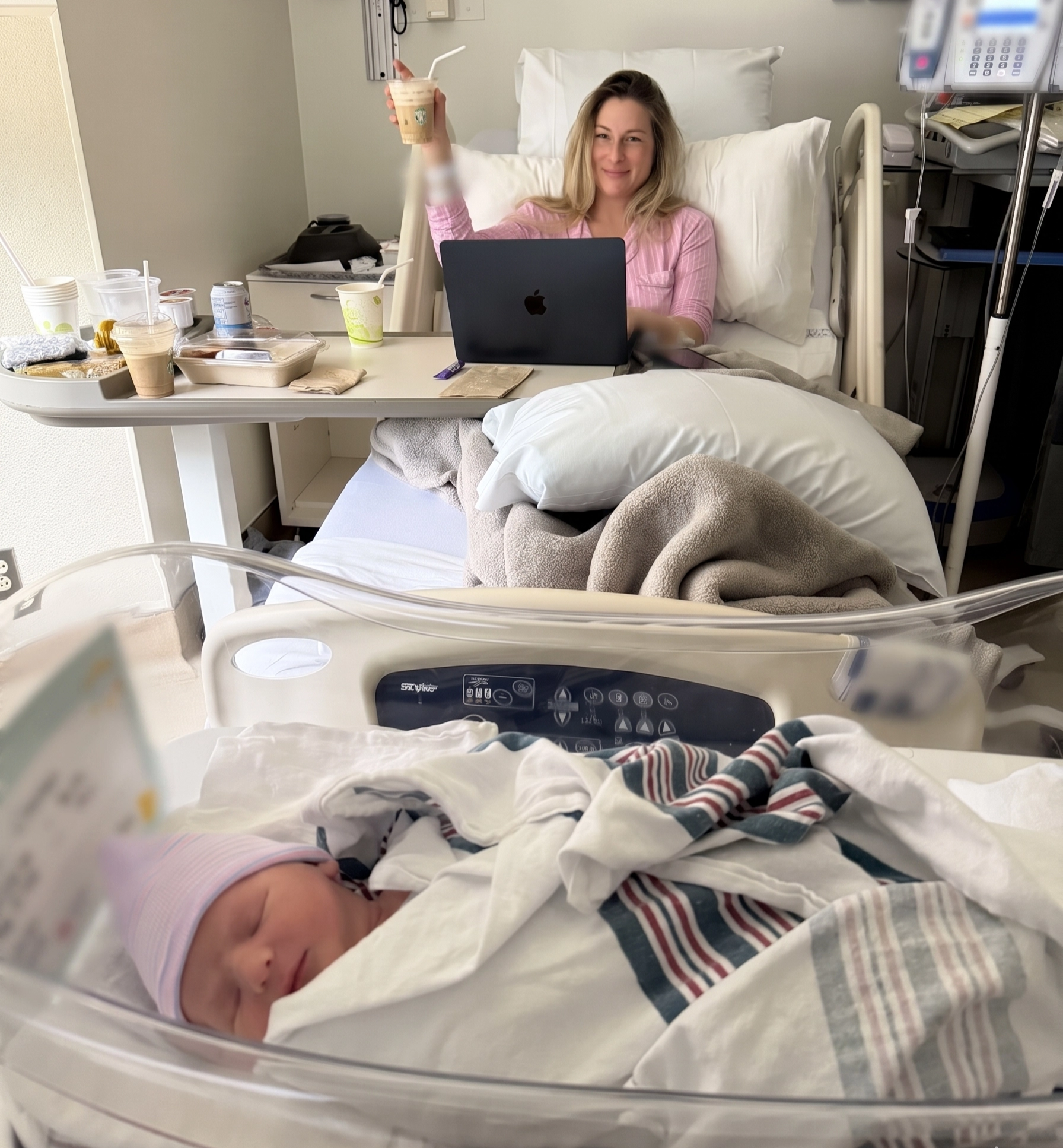 Woman in a hospital bed raising an iced coffee, working on a laptop, with a newborn asleep in a bassinet in the foreground