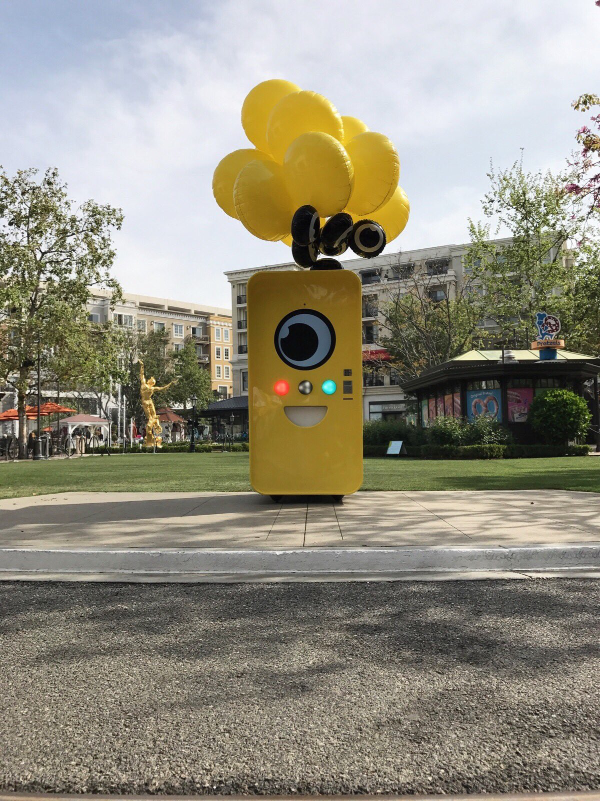 Yellow Snapbot Spectacles vending machine with a smiling camera-lens face and balloons in an outdoor shopping plaza