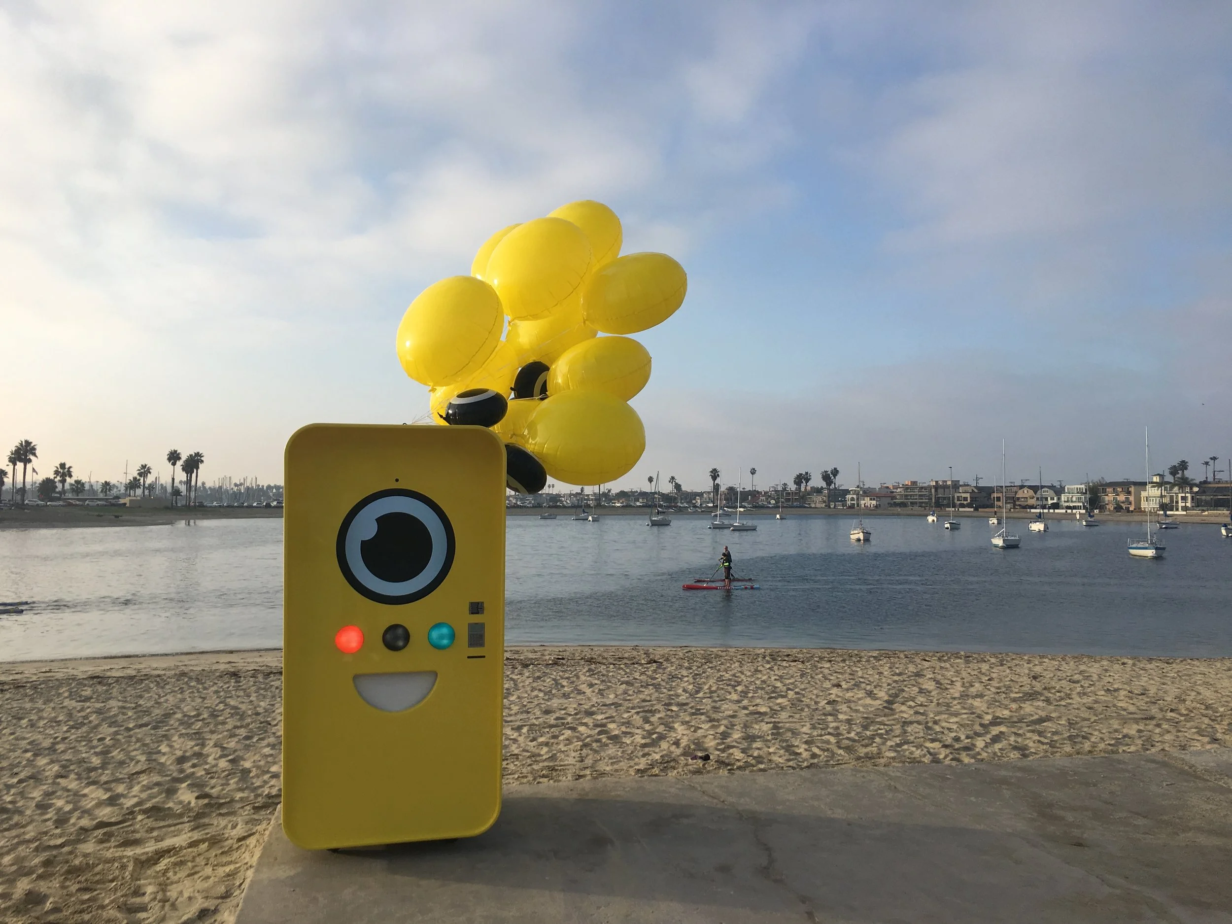 Yellow Snapbot vending machine with balloons on a beach; sailboats and a paddleboarder on the bay behind.