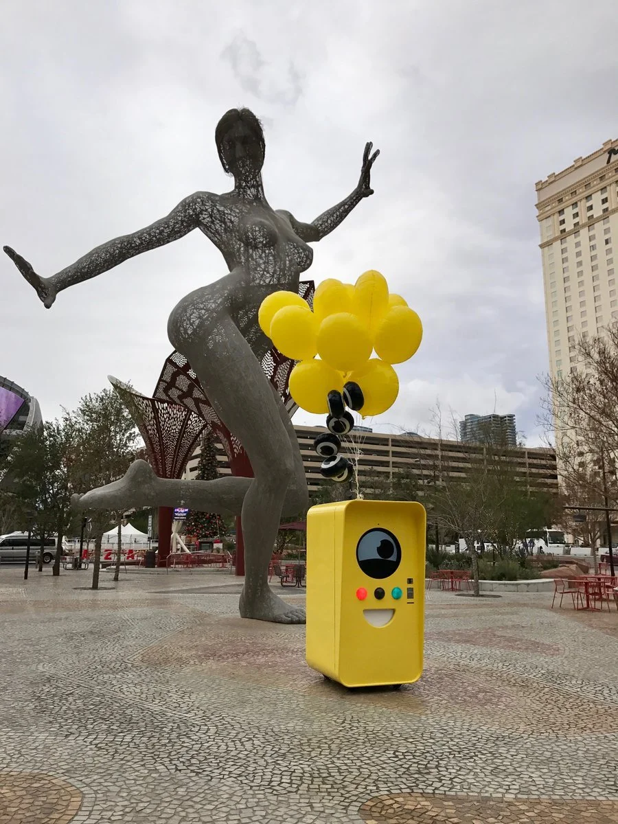 Yellow Snapbot Spectacles vending machine with balloons in a plaza before the Bliss Dance female wire-mesh statue.