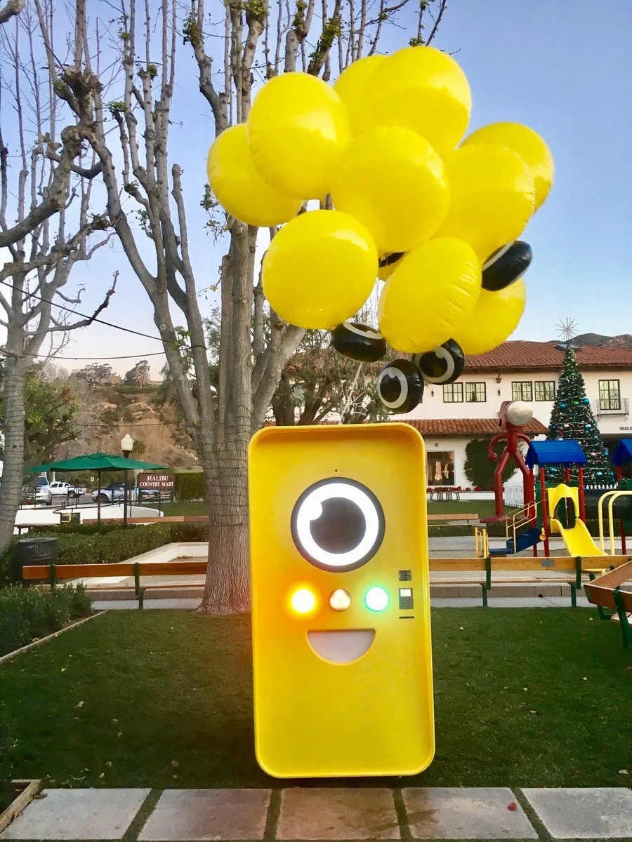 Yellow Snapbot Spectacles vending machine with balloons on a lawn beside a playground and Malibu Country Mart sign.
