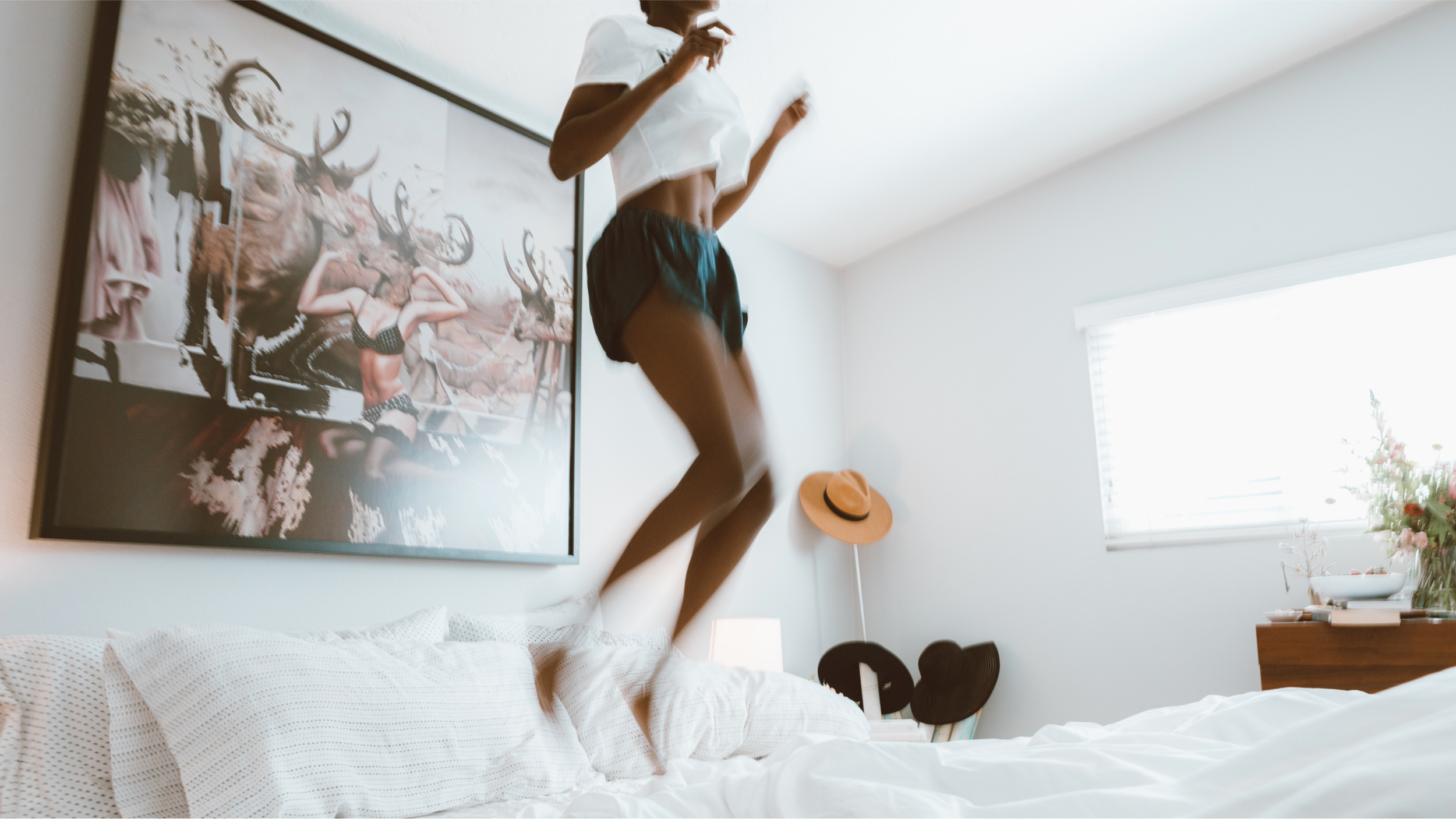 Woman in white top and shorts jumping on a bed beneath a large framed photograph in a bright bedroom