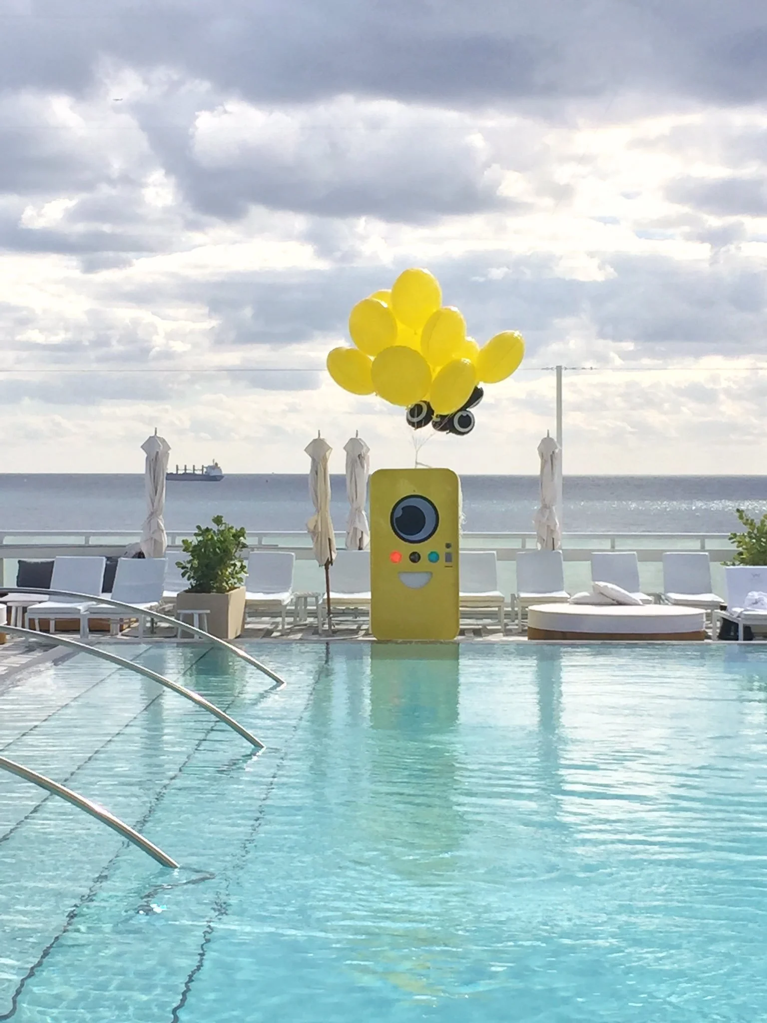 Yellow Snapbot vending machine with balloons standing at the edge of a poolside deck overlooking the ocean