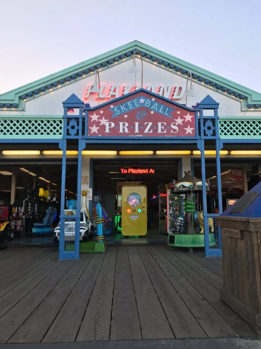 Boardwalk arcade with "Playland Skee-Ball & Prizes" sign; a yellow Snapbot vending machine glows in the entryway at dusk.