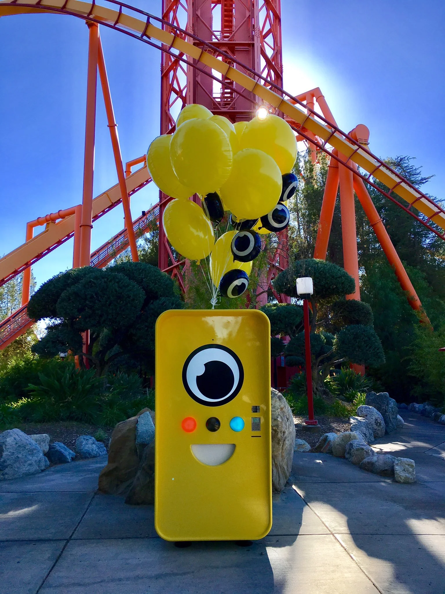 Yellow Snapbot Spectacles vending machine with balloons beneath a tall red roller coaster structure at a theme park.