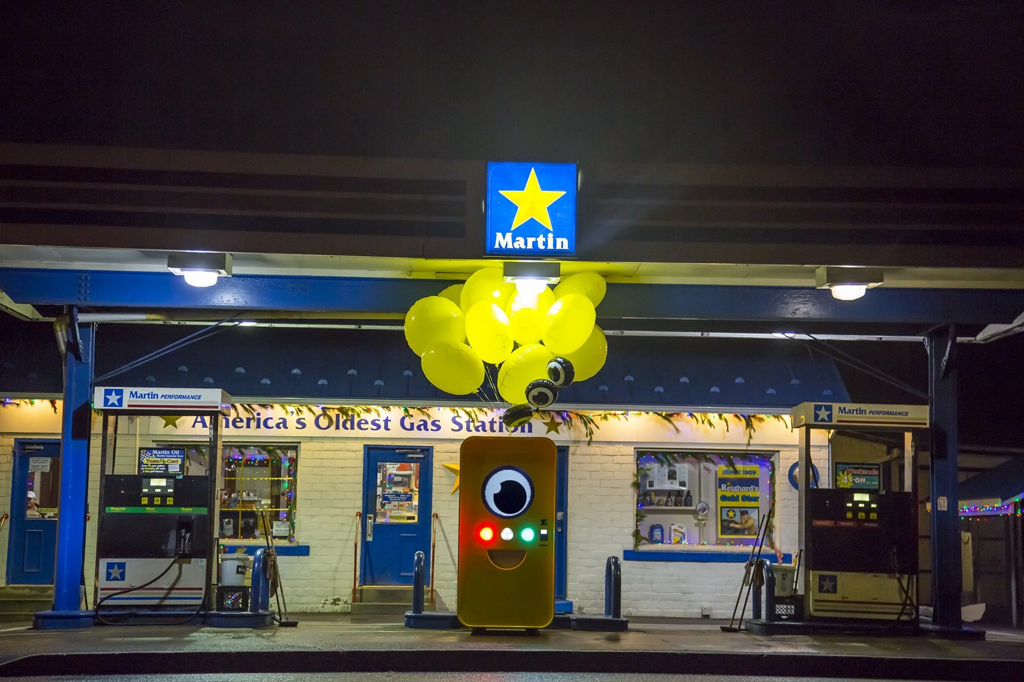 Yellow Snapbot Spectacles machine with balloons at night under a Martin gas station sign reading "America's Oldest Gas Station".