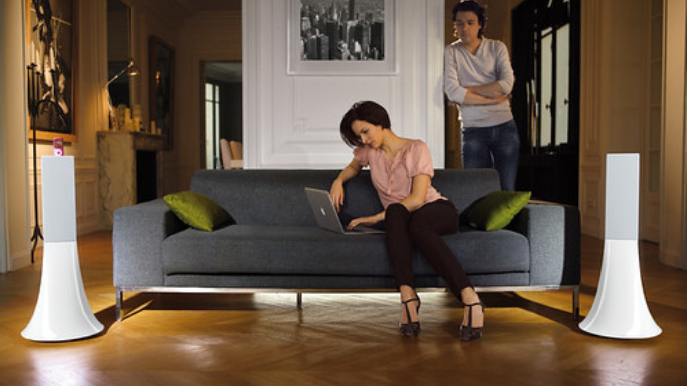 Two tall white Zikmu tower speakers flanking a sofa where a woman uses a laptop, another woman standing