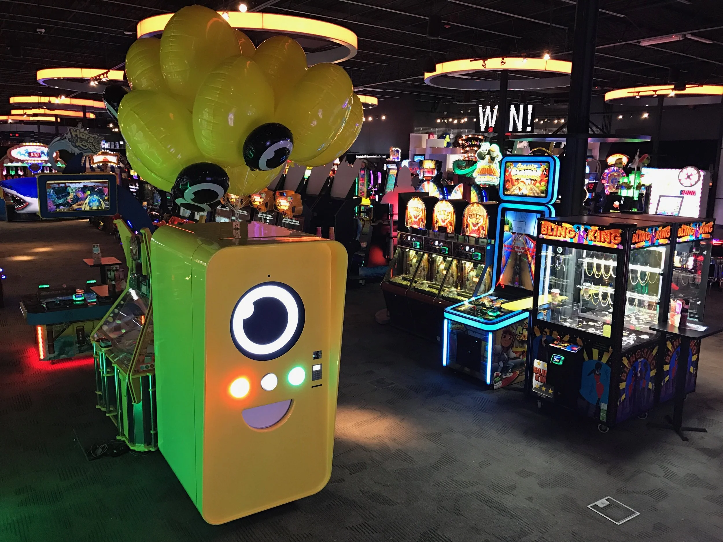 Yellow-green Snapbot Spectacles vending machine with balloons inside an arcade of claw and coin games.