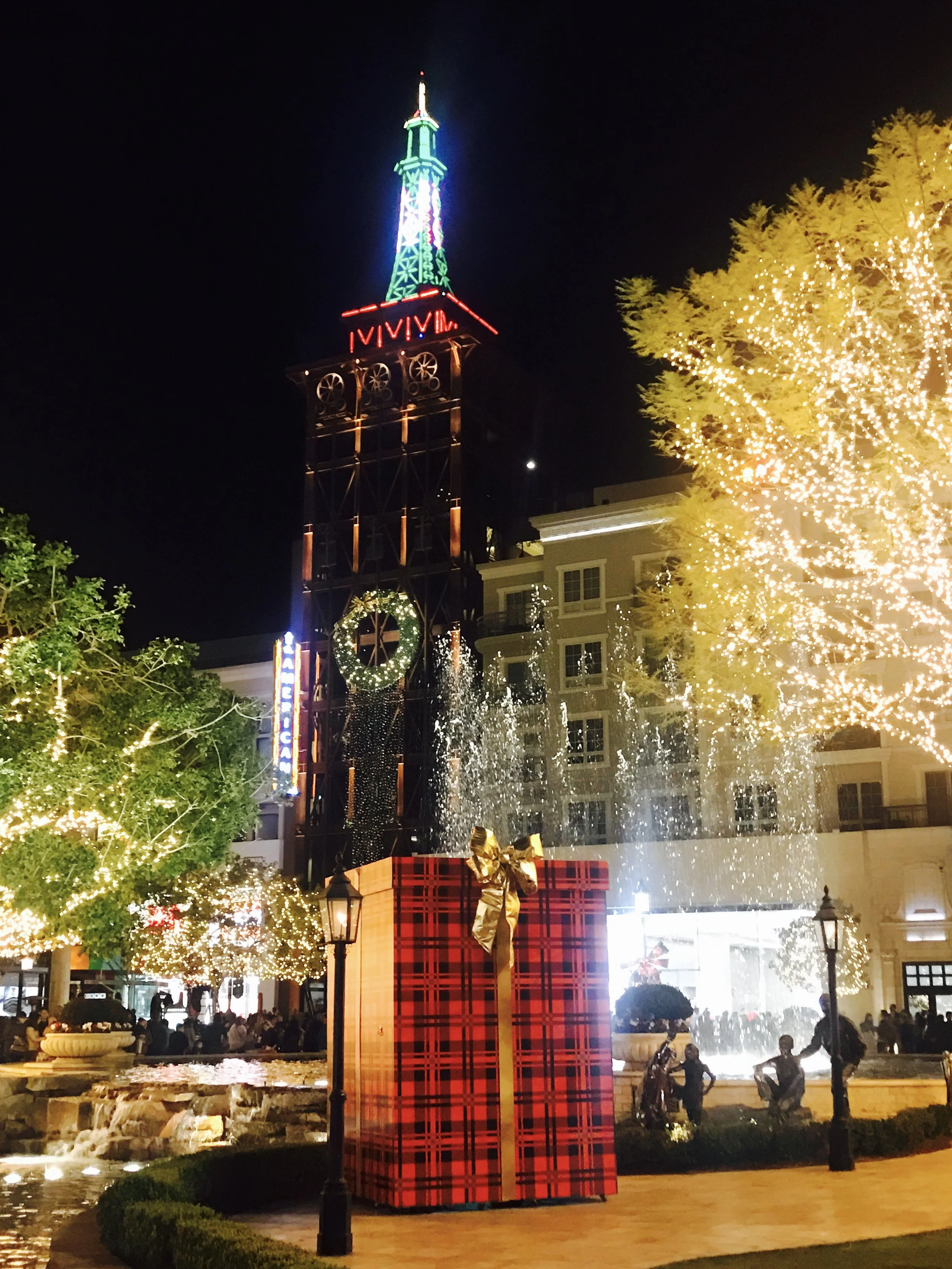 Nighttime shopping plaza with a lit Eiffel-style clock tower, sparkling trees, fountain and a giant plaid gift box.
