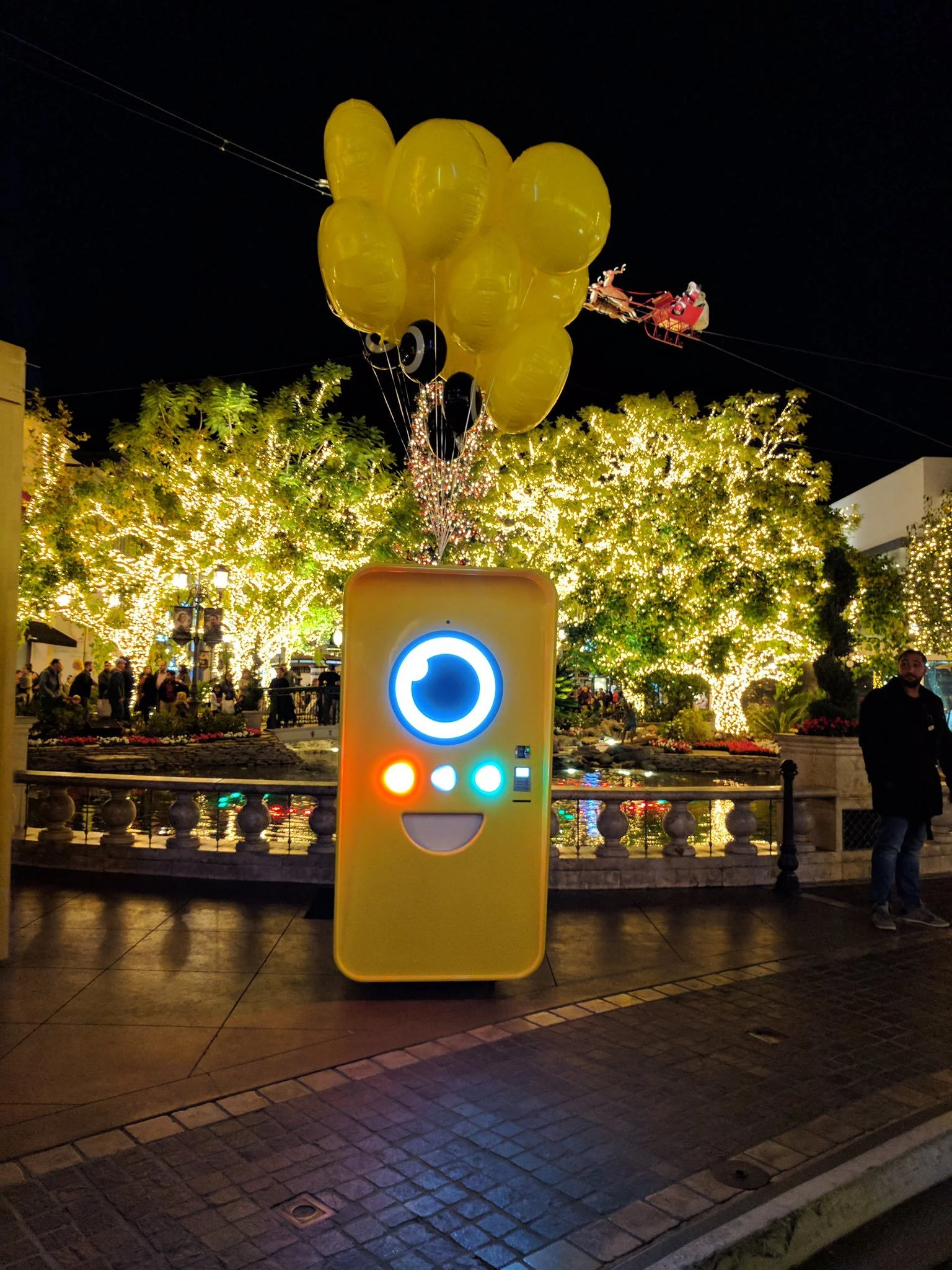 Yellow Snapbot Spectacles machine with glowing face and balloons at night, before tree lights and a Santa sleigh zip-line.