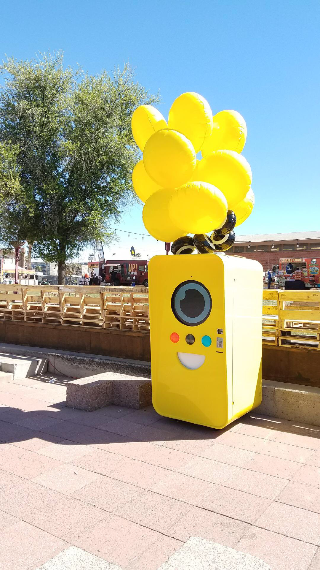 Yellow Snapbot Spectacles vending machine with balloons beside empty bleachers and food trucks under blue sky