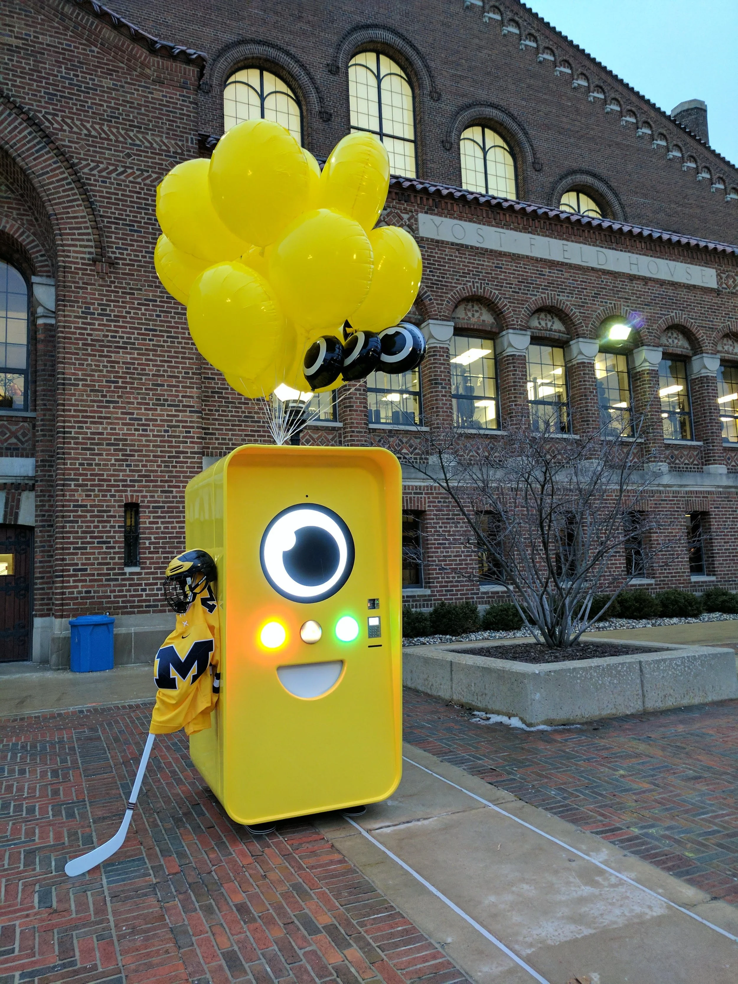 Yellow Snapbot vending machine with balloons outside Yost Field House; a person in Michigan hockey gear beside it.