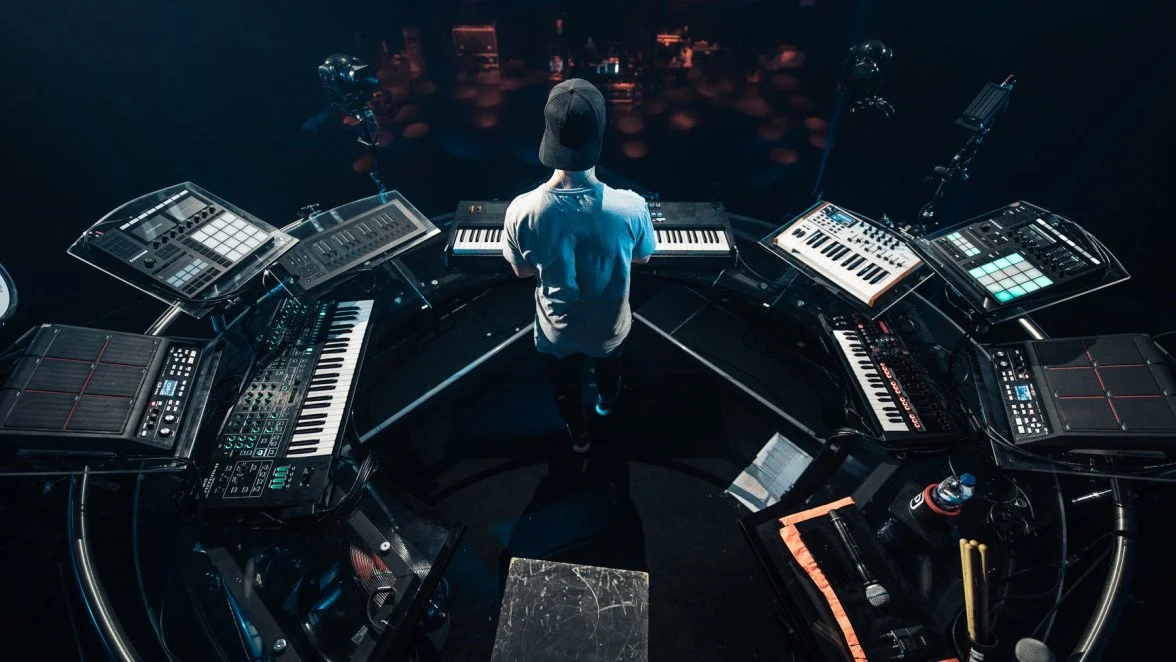 Overhead shot of a performer in a cap seen from behind, ringed by keyboards, synths, and drum pads on a dark stage.