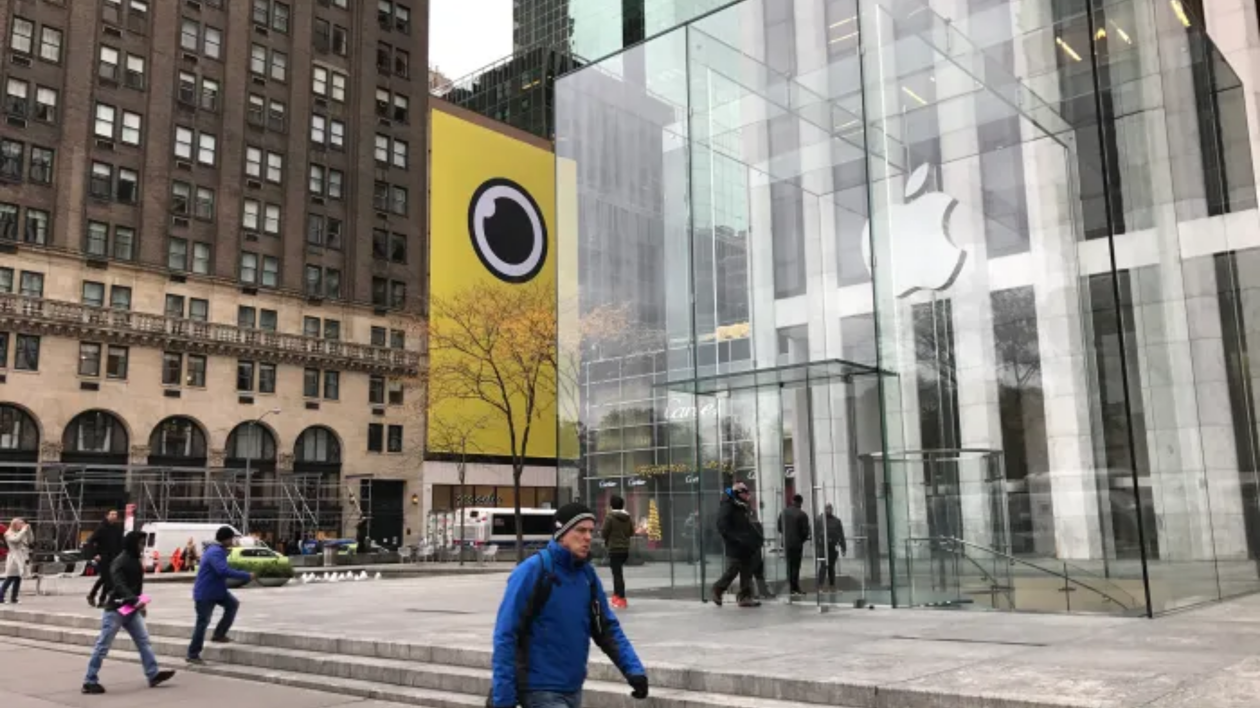 Fifth Avenue glass-cube Apple Store beside a yellow Snapchat Spectacles billboard; pedestrians walk by