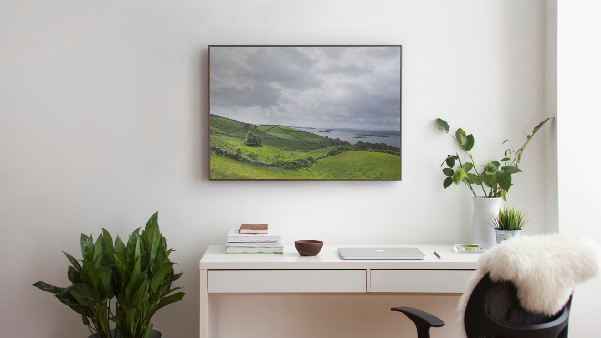 Framed green landscape photo of rolling hills and a lake hung above a white desk with a laptop and plants