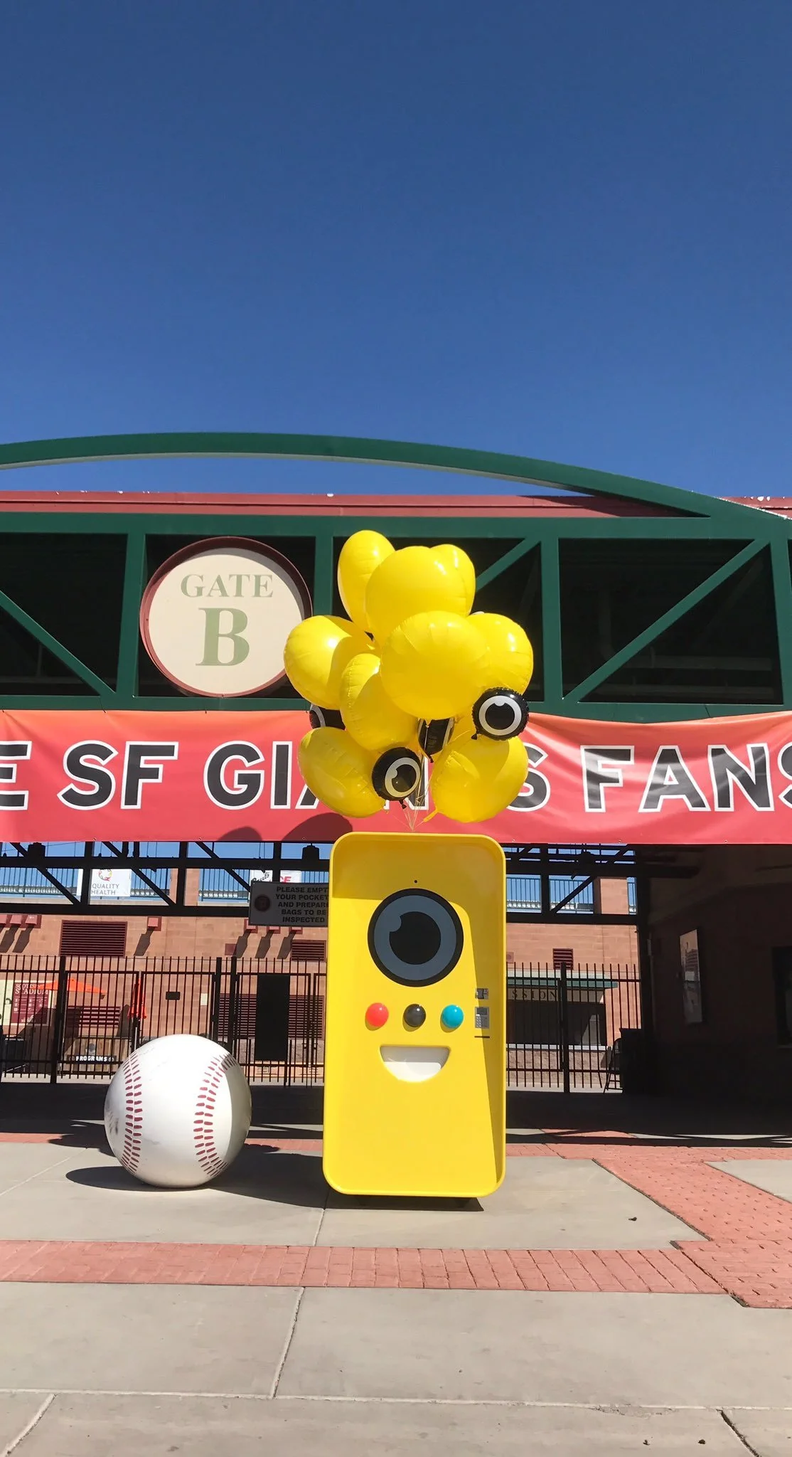 Yellow Snapbot vending machine and balloons beside a giant baseball at a Gate B entrance with an SF Giants fans banner