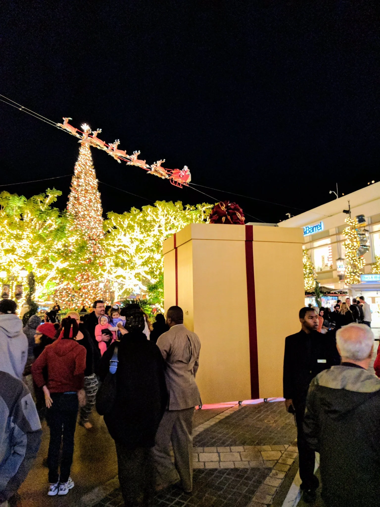 Nighttime crowd at an outdoor mall by a lit Christmas tree and a giant gift-box structure, Santa sleigh strung overhead.