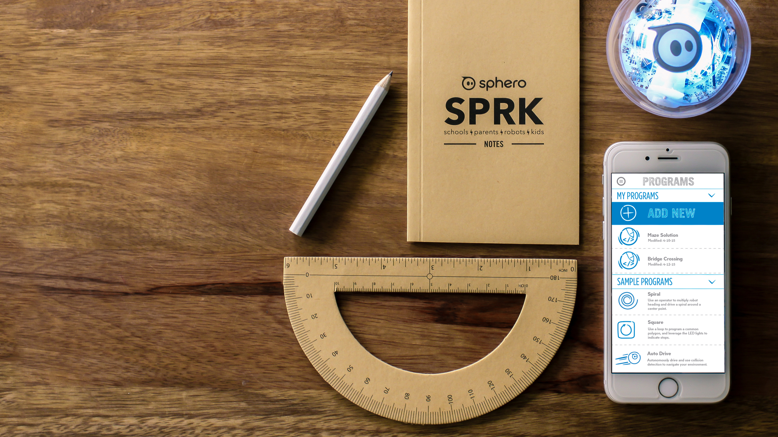 Sphero SPRK+ clear robot ball, kraft SPRK notebook, protractor and phone showing the Programs app on wood