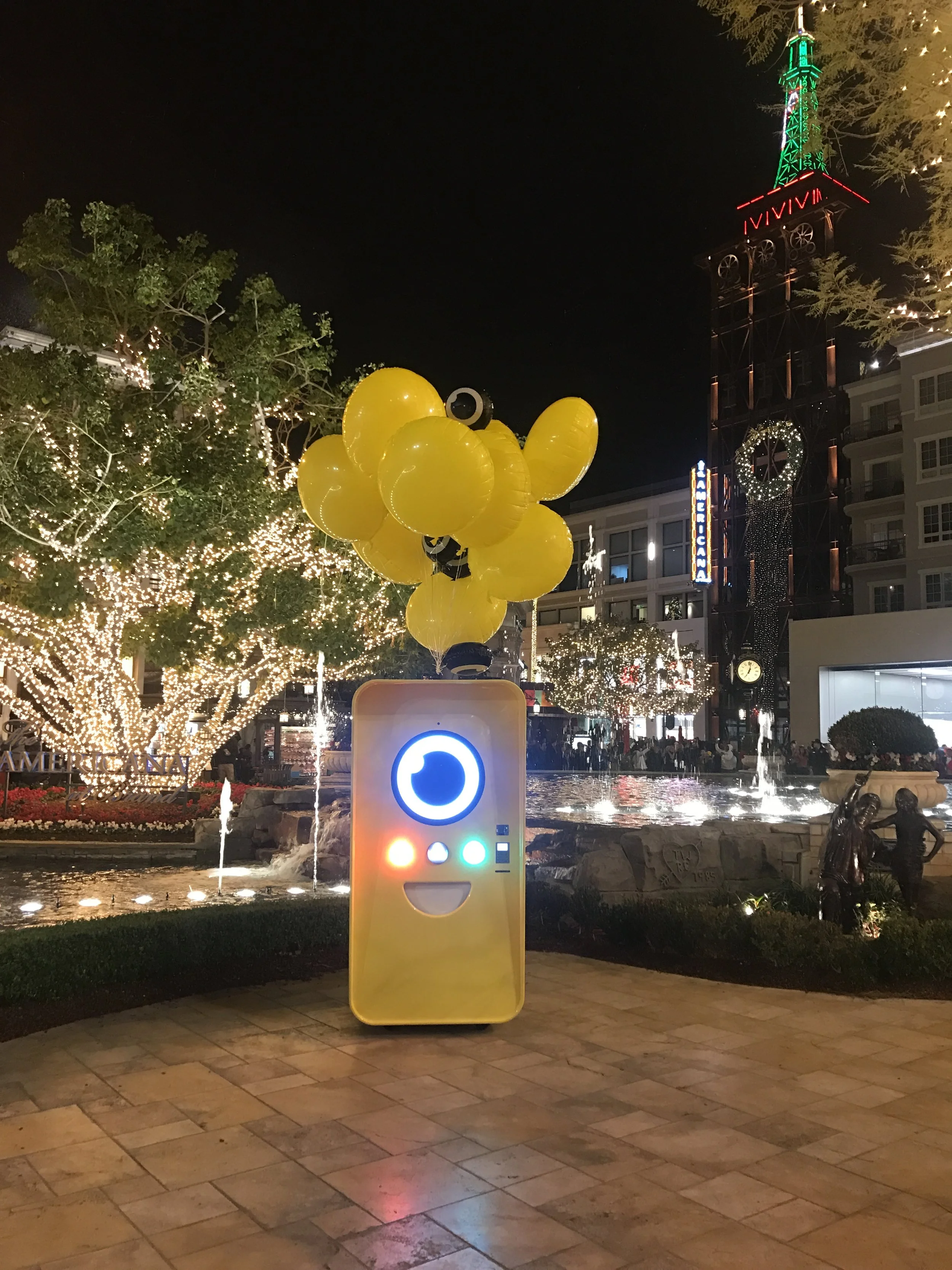 Yellow Snapbot Spectacles vending machine with balloons at night, lit trees and Americana clock tower behind it.