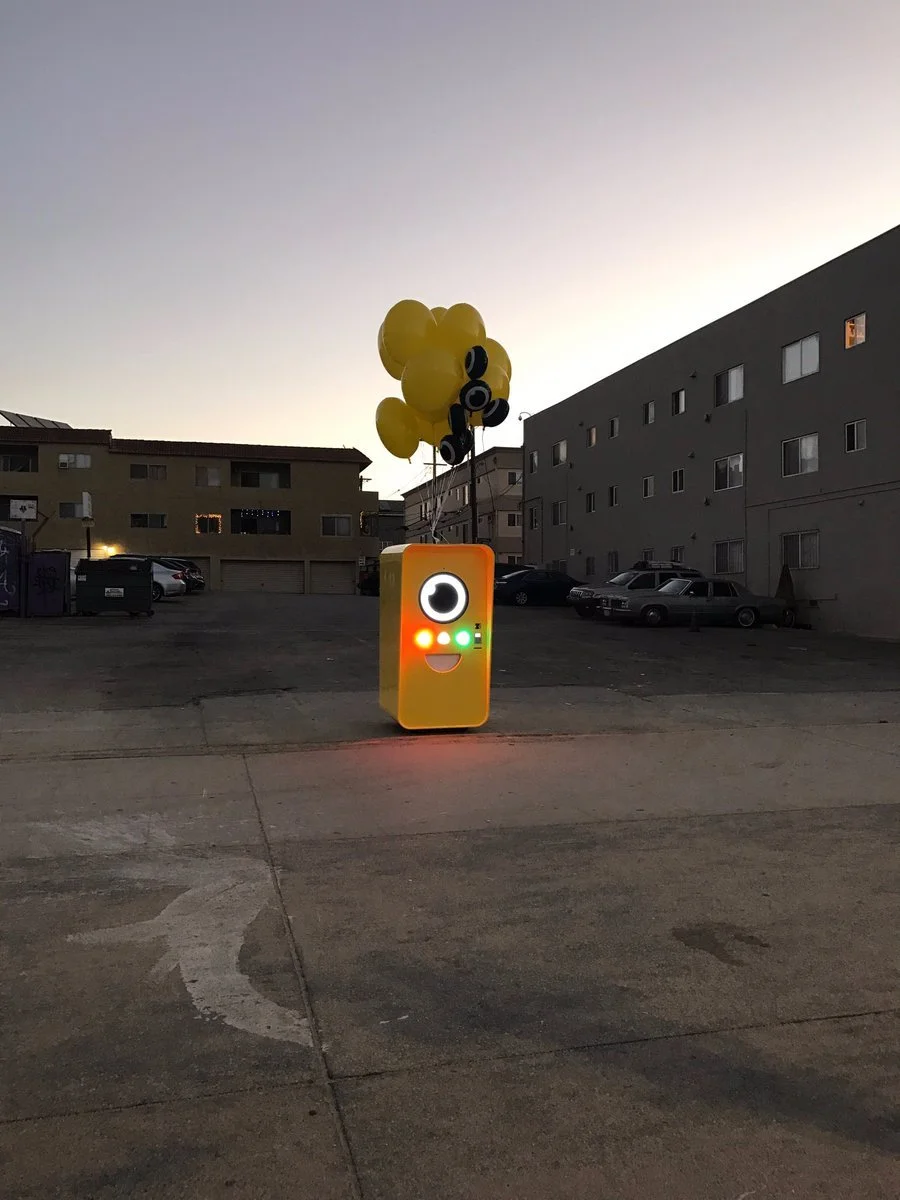 Orange-yellow Snapbot vending machine tethered to yellow balloons in a parking lot at dusk