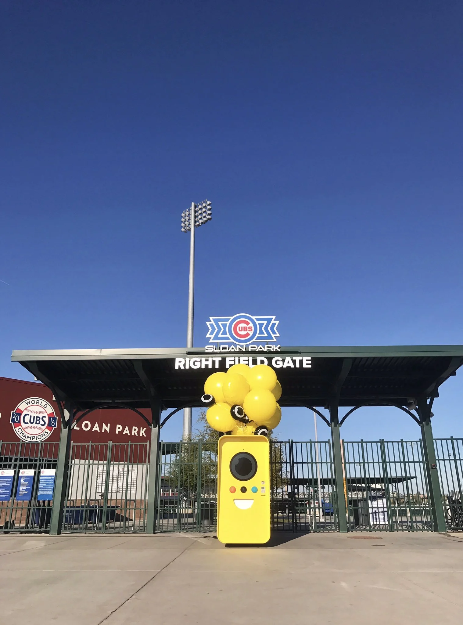 Yellow Snapbot vending machine with balloons at the Cubs Sloan Park Right Field Gate under a clear blue sky