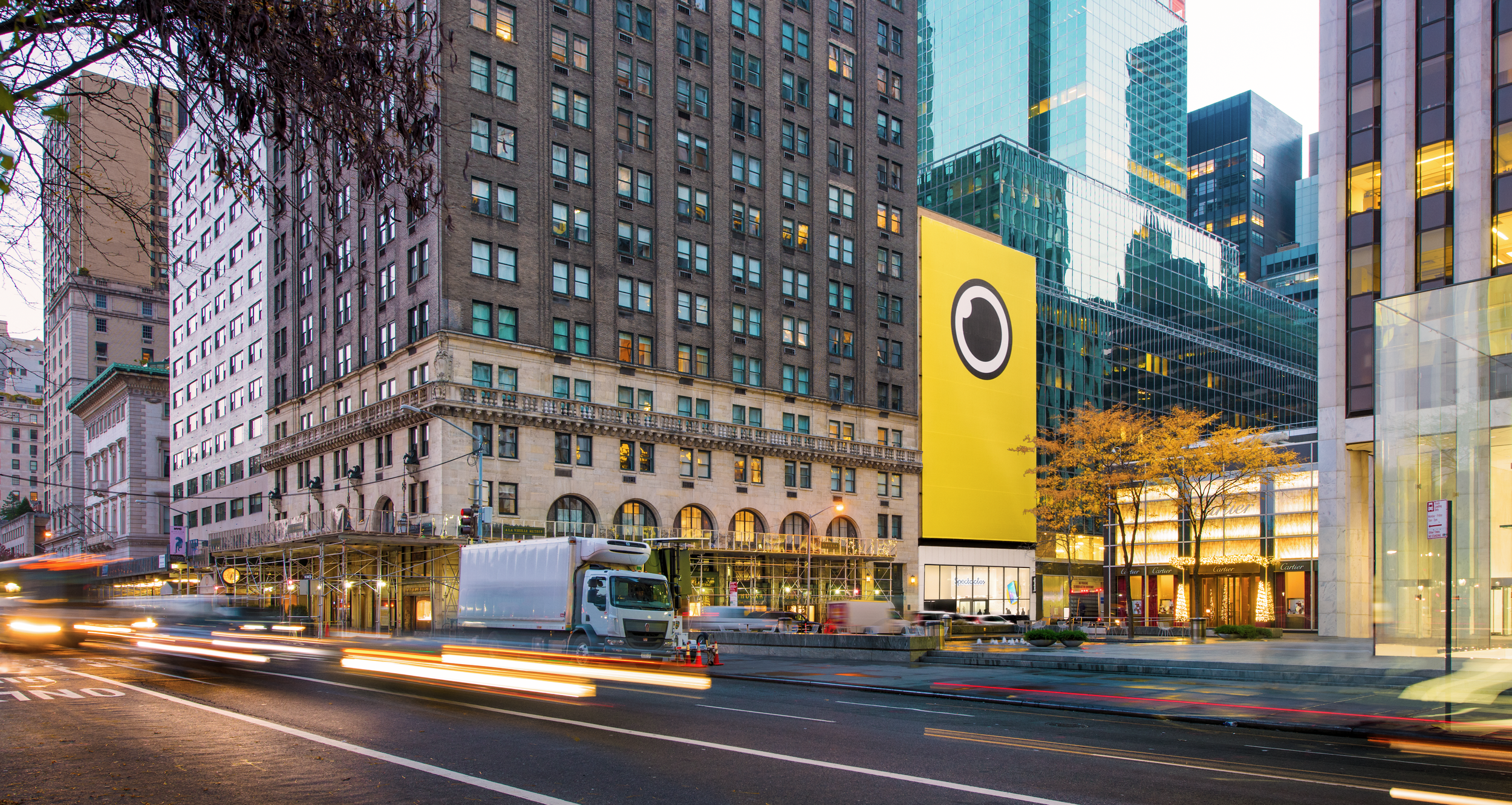 City street at dusk with a large yellow Snapchat Spectacles ghost logo billboard on a building near Cartier