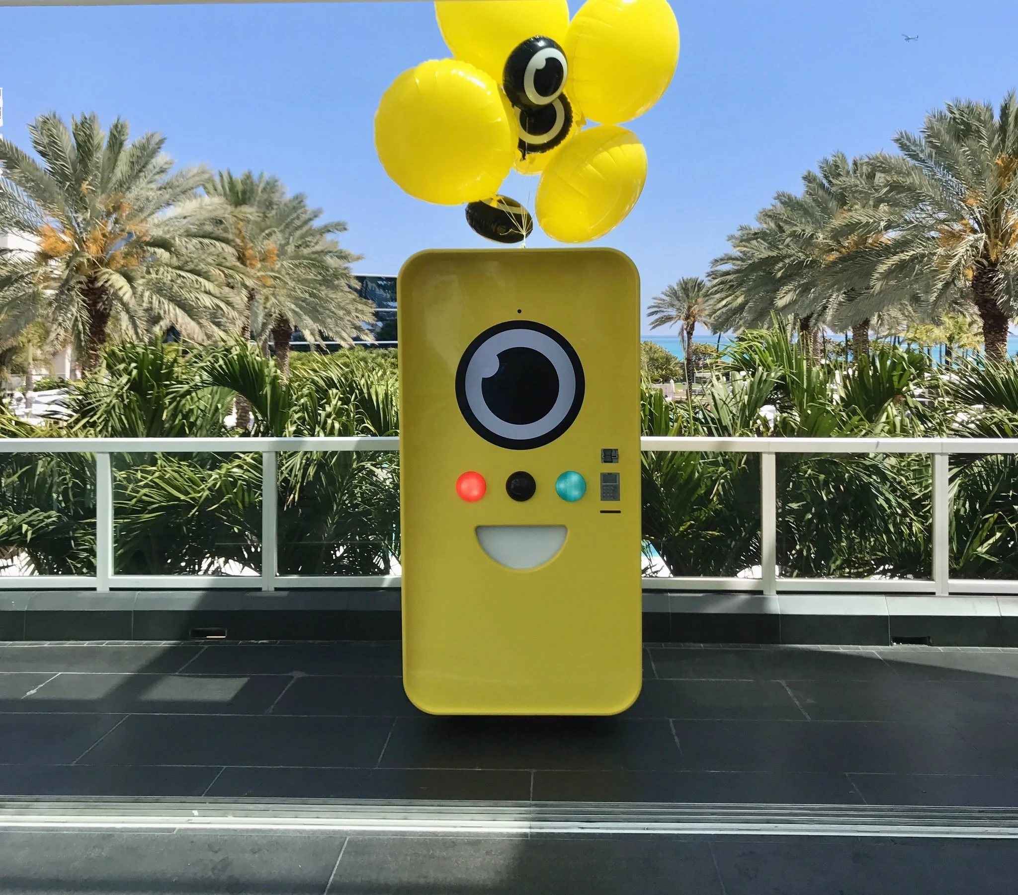 Yellow Snapbot vending robot with camera-eye face and balloons on a poolside deck ringed by palm trees